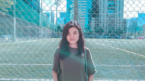 Portrait of beautiful young woman standing by chainlink fence