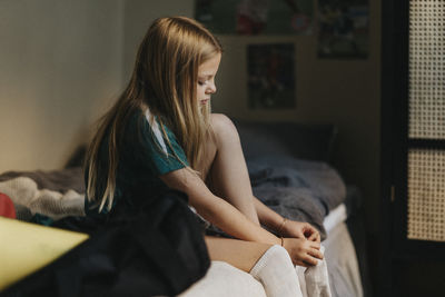 Blond elementary girl wearing sock while sitting on bed at home