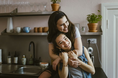 Portrait of happy woman embracing female friend from behind while sitting in kitchen at home