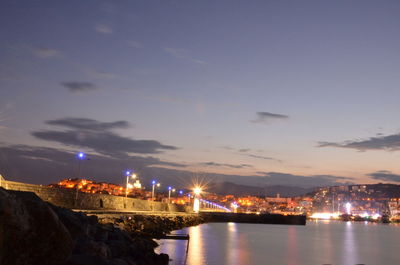 Illuminated buildings by sea against sky at night