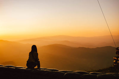 Rear view of man sitting on mountain against orange sky