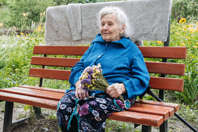 Senior elderly woman with gift, flowers bouquet and basket of groceries sits on bench outdoor