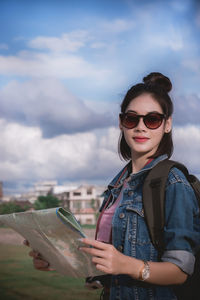 Portrait of young woman wearing sunglasses while holding map against sky