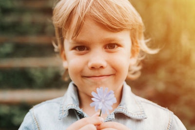 Portrait happy dishevelled long blond hair preschool kid boy holding delicate pale lilac wildflower