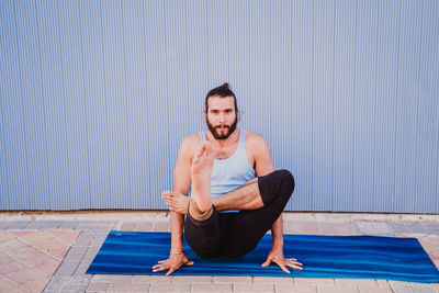 Full length of man performing yoga on exercising mat against wall