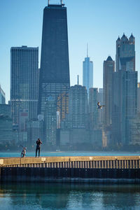 Man photographing with camera while standing on retaining wall by lake with city buildings seen in background