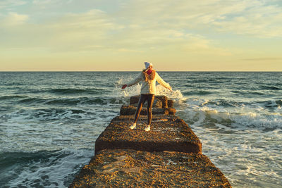 A woman with blond hair in a hat and a fur coat walks along the sea along a stone breakwater