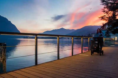 Woman with baby stroller walking by lake on pier during sunset