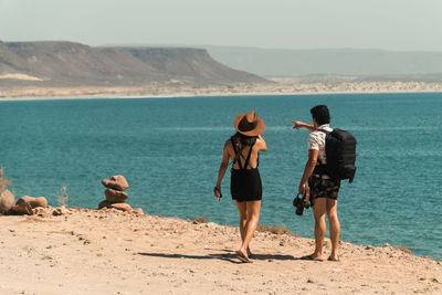 A male and female photographer couple stand on cliffside overlooking the blue sea in mexico.