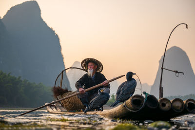Man sitting on fishing rod against sky during sunset