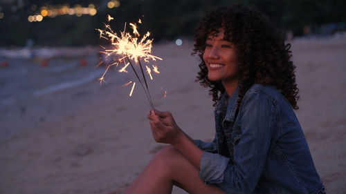 Smiling young woman playing with sparkler while standing on beach