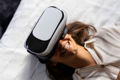 From above happy young female in vr headset smiling while lying on bed and exploring cyberspace at home