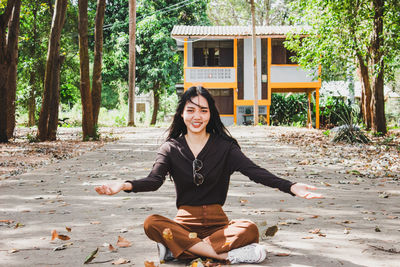 Portrait of smiling young woman throwing leaves while sitting on road in forest