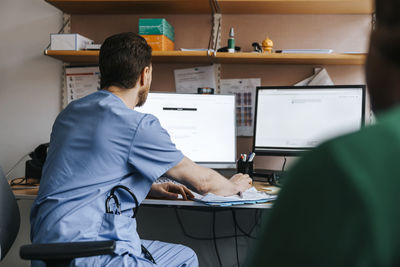 Male physician in blue scrubs checking medical records on computer while sitting in examination room at hospital