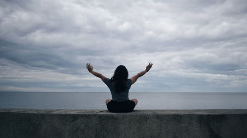 Rear view of woman sitting with arms outstretched on retaining wall against sea