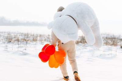 Rear view of man carrying stuffed toy with balloons on snow covered land