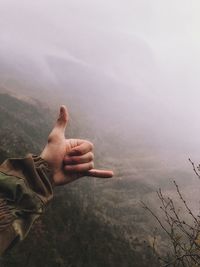 Close-up of person doing shaka sign on mountain during foggy weather