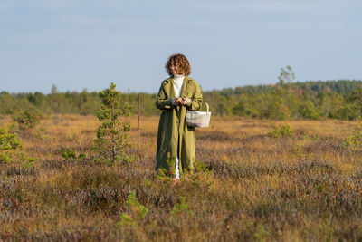 Happy lady in trench coat looks for cranberries among grass resting in autumn nature