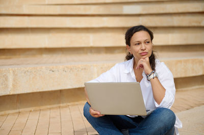 Young woman using laptop while sitting against wall