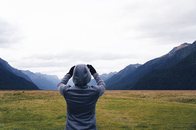 Rear view of person in hooded jacket photographing mountains against sky