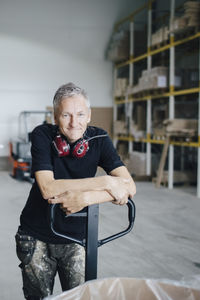 Portrait of mature worker leaning on pallet jack by stack at industry