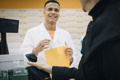 Midsection of senior female customer using contactless payment through smart phone against pharmacist at checkout in sto