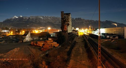 Railroad tracks by factory against mountains at night