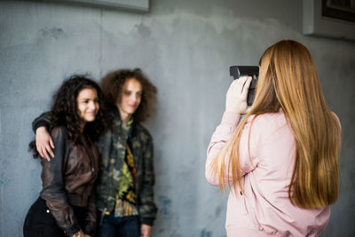 Rear view of girl photographing teenage friends standing against wall at parking garage