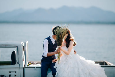 Couple kissing while sitting on car by sea against sky