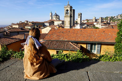 Back view of girl sitting on wall in upper city enjoying cityscape of bergamo, lombardy, italy.