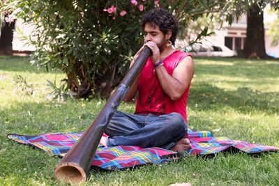 Man playing traditional wind instrument while sitting on throw rug in park