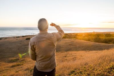 Rear view of man shielding eyes at shore against sky during sunset