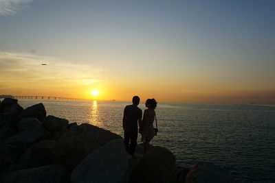 People on rocks by sea against sky during sunset