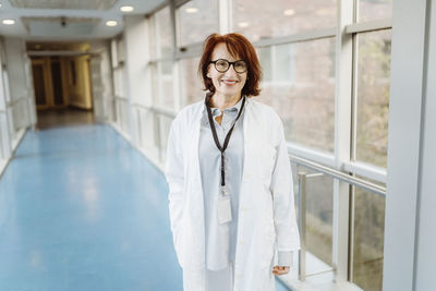 Portrait of smiling senior female doctor wearing id card and lab coat standing in hospital corridor