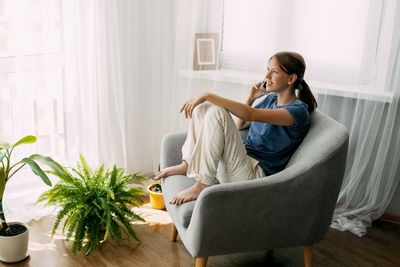 A cheerful teenage girl is sitting in an armchair and talking on the phone at home