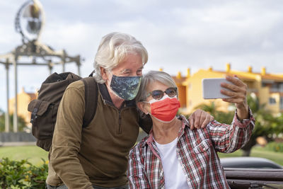 Cheerful senior couple wearing mask doing selfie while standing outdoors