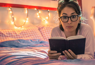Young woman reading book on bed at illuminated home 