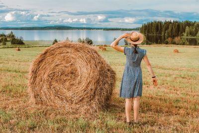 A girl in a dress and a straw hat stands with her back to the hay field. summer landscape