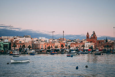 Sailboats moored on sea by buildings against sky during sunset