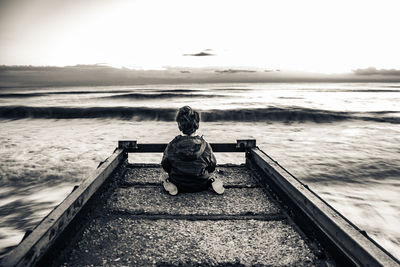 Rear view of boy sitting on jetty against sea