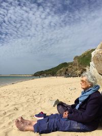 Side view of senior woman sitting at beach against sky