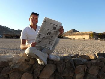 Full length portrait of smiling man with newspaper sitting on stone wall at beach