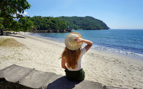 Holidays in brazil. back view of traveler girl sitting on wall in ubatuba beach, brazil.