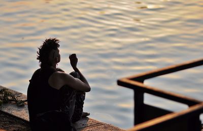 Rear view of woman with cigarette sitting on retaining wall by river