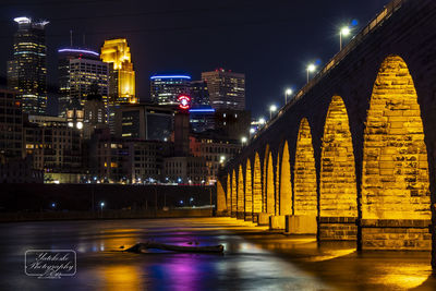 Illuminated bridge over river by buildings against sky at night