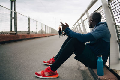 Sportsman holding mobile phone while looking at female athlete running on footbridge