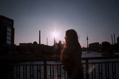 Side view of woman standing on railing against sky during sunset