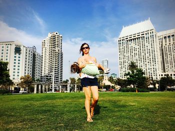 Portrait of mother wearing sunglasses carrying daughter while walking on grassy field against blue sky during sunny day