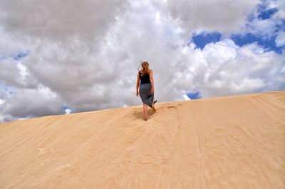 Rear view of woman walking on sand dune against cloudy sky