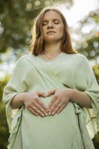 Happy pregnant woman makes love heart sign on her round belly. closed eyes, green trees, sunlight 